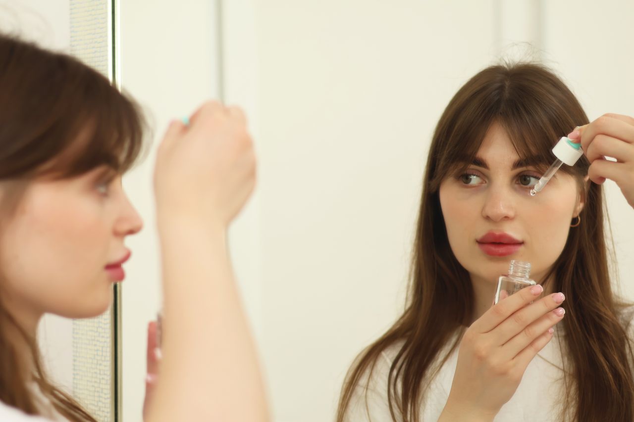 A young woman applying skincare serum in front of a mirror indoors. Perfect for beauty and self-care themes.
